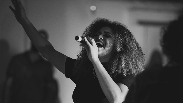 A woman with curly hair wearing a black tank top holding a mic and singing soulfully in a room with blurred people in the background.