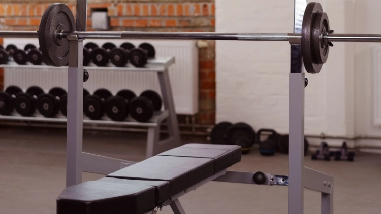 A barbell on bench press rack with dumbbell rack on the background.