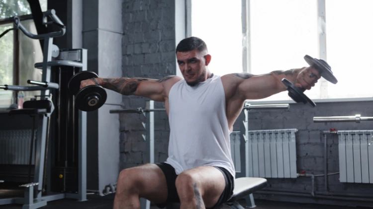 A big muscular male bodybuilder wearing a white tank top and black shorts seating on a bench while performing a dumbbell lateral raise exercise in a gym with concrete wall and windows in the background.
