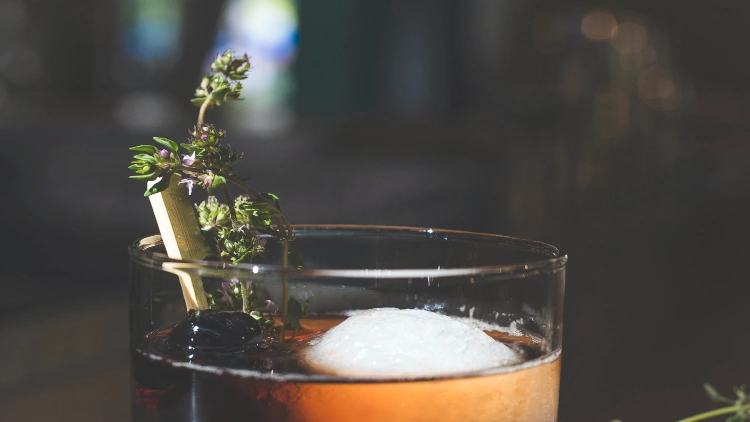 An alcoholic cocktail in a short brown glass filled with ice and garnished with a sprig of fresh rosemary.