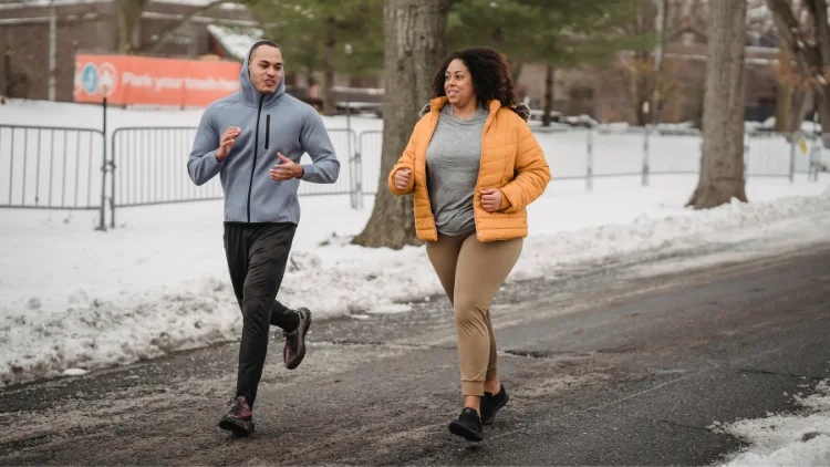 A couple jogging outdoors in the winter, they are both wearing warm athletic clothing, including jackets, leggings, snow is visible on the ground, and trees are visible in the background, the couple is shown in motion, with their legs mid-stride as they jog together in the scenic winter landscape.