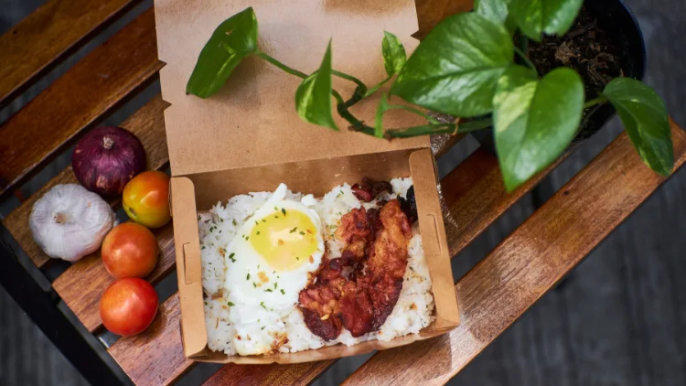 Delicious meal in a box with a generous serving of steaming white rice topped with a sunny-side-up egg and crispy bacon strips, tomatoes, garlic, and onions can be seen, adding a burst of color and flavor to the meal.