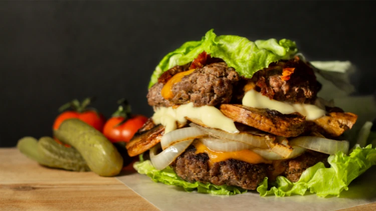 A mouth-watering lettuce wrapped burger with two juicy beef patties topped with melted cheese and sliced onions, the burger is garnished with a crisp pickle and fresh tomato in the background, adding a pop of color to the dish, this low-carb and high-protein meal option is perfect for those looking for a healthier yet satisfying alternative to traditional burgers.