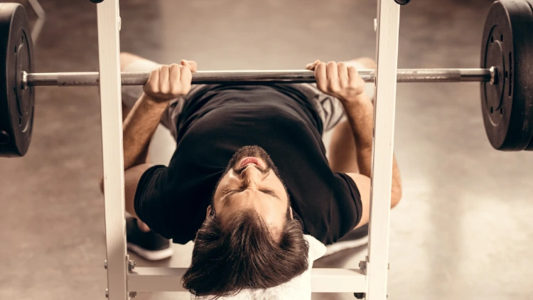 A man with beard is lying on a white bench with a barbell above him, doing a close grip bench press exercise, he has his hands close together on the barbell, and is using his triceps and chest muscles to lower and raise the weight.