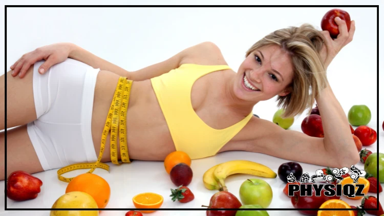 A blonde woman is laying on her side in a yellow top and white shorts with a measuring tape around her waist behind assorted fruits such as apples, bananas oranges, and strawberries while smiling due to her results on a holistic weight loss plan.