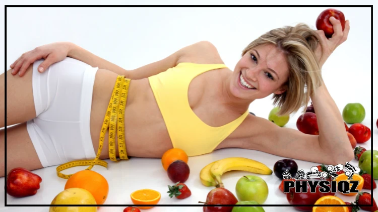 Blonde woman in yellow top and white shorts with measuring tape around waist, surrounded by apples, bananas, oranges, and strawberries, smiling from the success of the holistic approach to losing weight.