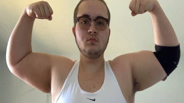 A man wearing glasses, flexing his arm muscle to show his progress to a skinny arm, the man's arm is visible in the foreground of the image, appearing slender with little muscle.