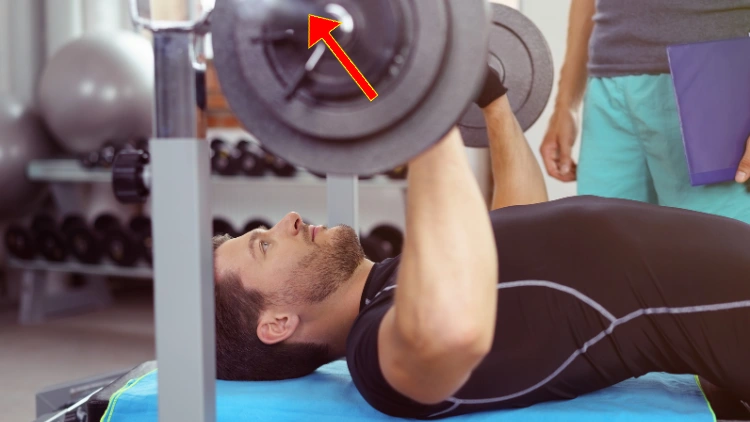 A bearded man wearing a black activewear shirt is performing a barbell bench press exercise, a red arrow can be seen as an indicator of the bench press path.