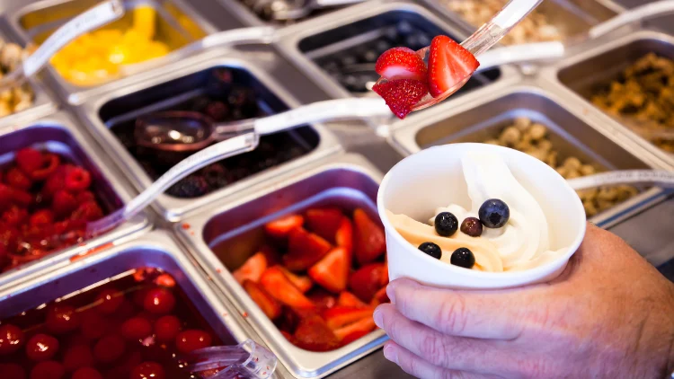 A colorful array of toppings neatly arranged on the countertop of a frozen yogurt store, including fresh fruits, a workers hand is seen putting fruits toppings on a yogurt.