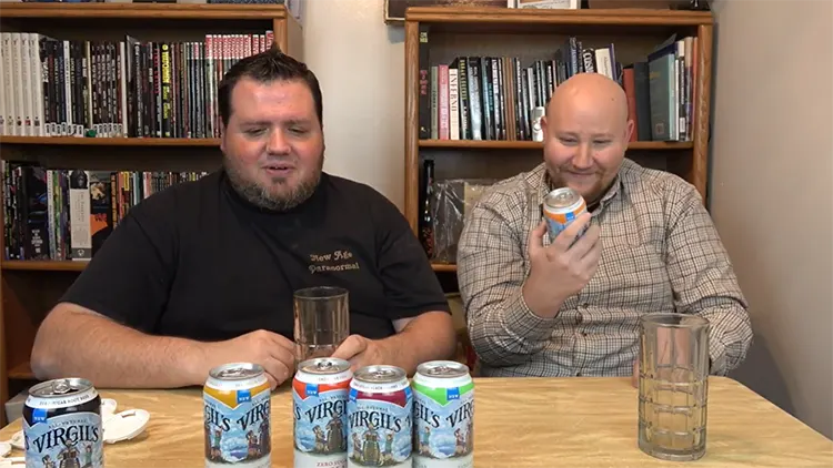 Two big men, one wearing a black t-shirt, and the other one wearing a flannel is checking different flavors of Virgil's zero sugar soda in a room with bookshelves in the background.
