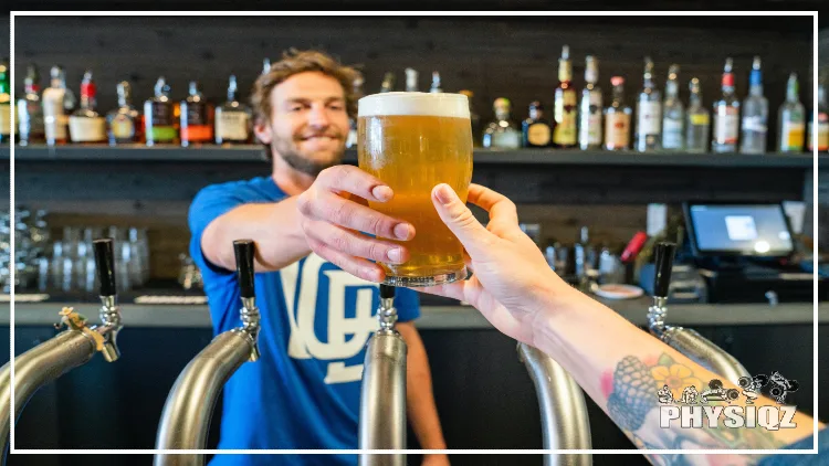 A smiling man, wearing a blue t-shirt with a white indistinguishable baseball logo is holding out a glass of beer towards the other person who is reaching to take the beer and questions why they're a few pounds less the day after drinking, and the person taking the beer has a tattooed arm and there is a shelf of liquor bottles in the background.