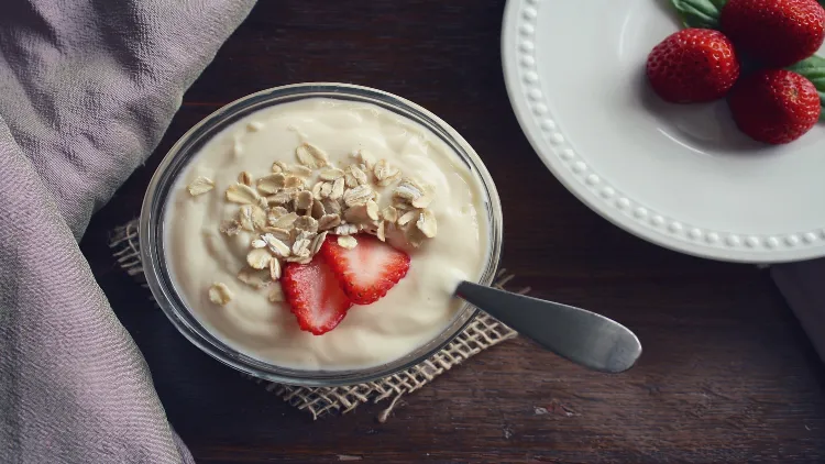 A bowl of yogurt with sliced strawberries arranged on top, the yogurt is white and appears creamy, while the strawberries are bright red and fresh, in the background, there is a plate of whole strawberries, some with leaves still attached.