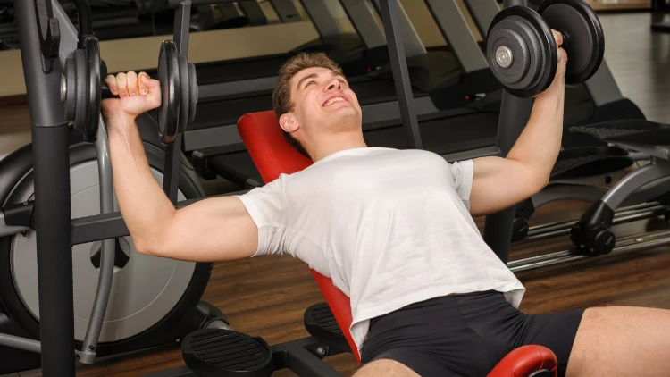 A young man wearing a white t-shirt and black shorts is performing an inclined bench press exercise using dumbbells in a gym with wooden floor.