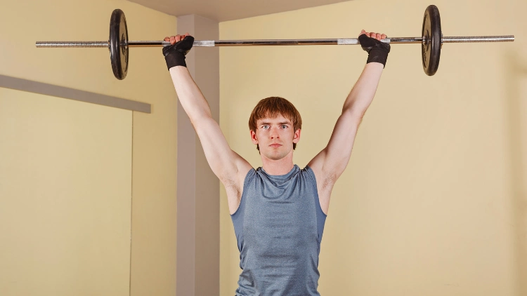 A young man with black gloves, wearing a grey tank top and black shorts is performing a standing overhead shoulder barbell press in a studio with light orange walls.