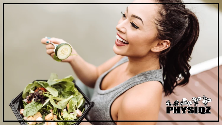 A fit woman with a ponytail hair wearing a gray top is holding a takeaway salad from Zaxby's gluten-free menu that is filled with fresh greens and a variety of colorful veggies, including a sliced cucumber.