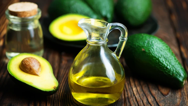 A clear jar filled with avocado oil placed on top of a wooden surface along with a whole and sliced fruit of avocado, another jar can also be seen in the background.