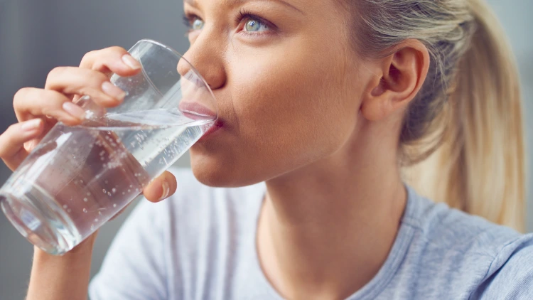 A blonde woman with grey-colored eyes, wearing a grey t-shirt is drinking water from a clear glass.