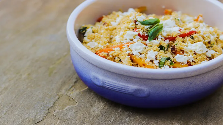 A blue bowl filled with food that is made with pepper, couscous, cheese, feta, basil, and rice, displayed on top of a wooden surface.