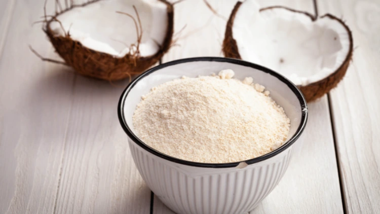 A white ceramic bowl filled with coconut flour and behind it in the background is a coconut shell broken in two pieces, and displayed on top of a light wooden surface.