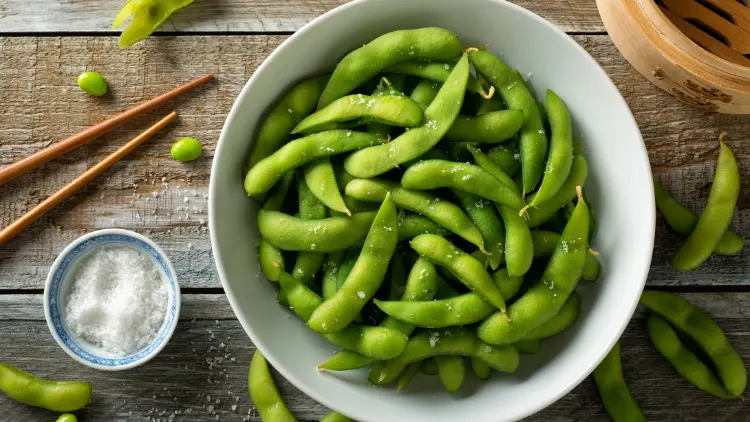A white bowl filled with long green bean vegetables called an edamame which can also be seen scattered around the table along with a cup of salt and a pair of chopsticks.