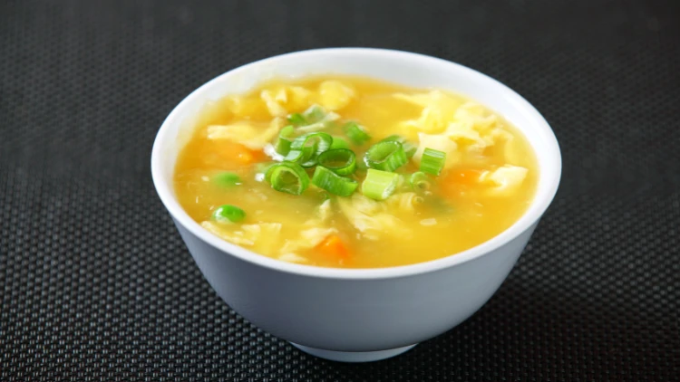 A white bowl filled with egg drop soup and garnished with minced scallions on top, and served on top of a black textile surface. 