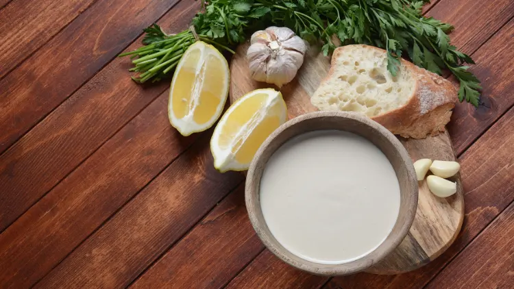 A wooden bowl filled with creamy tahini sauce, surrounded by slices of lemon, garlic, and fresh cilantro, a stack of sliced bread rests on the side of the bowl, the various ingredients suggest a recipe or serving suggestion, possibly for a savory dip or spread.