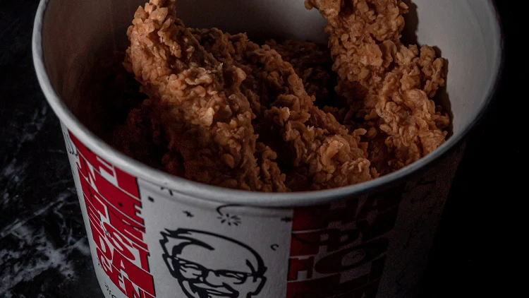A white bucket from KFC filled with fried chicken, placed in a dimly lit room.