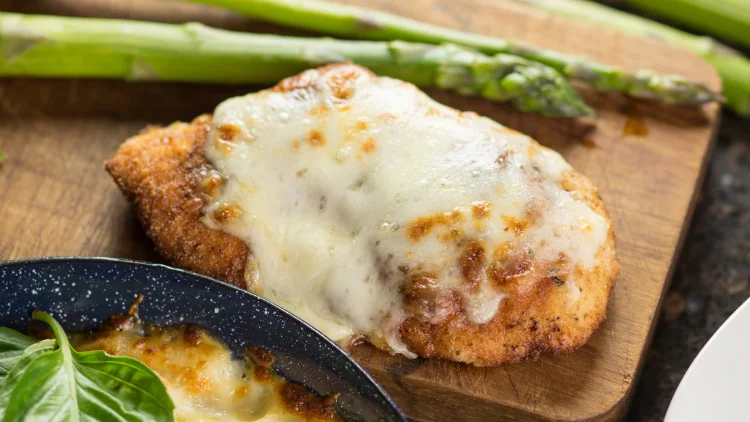On a wooden board is a piece of fried chicken with melted parmesan cheese on top, beside are a couple of asparagus, and a black bowl of parmesan cheese.