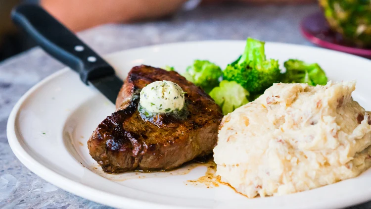 A deliciously prepared steak is featured on a white plate alongside a side of mashed potatoes and a stalk of broccoli, a pat of butter sits atop the steak, adding a rich and flavorful touch, a knife is placed near the steak on the plate, ready to be used.