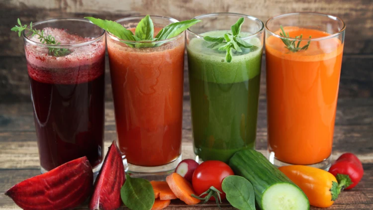 Four glasses filled with freshly made vegetable juices, each glass accompanied by a whole vegetable that was used to make the juice, the vegetables include a red beet, two carrots, a ripe tomato, and a green cucumber, the glasses are arranged neatly on a wooden table, with the colorful vegetables adding an aesthetic touch to the image.