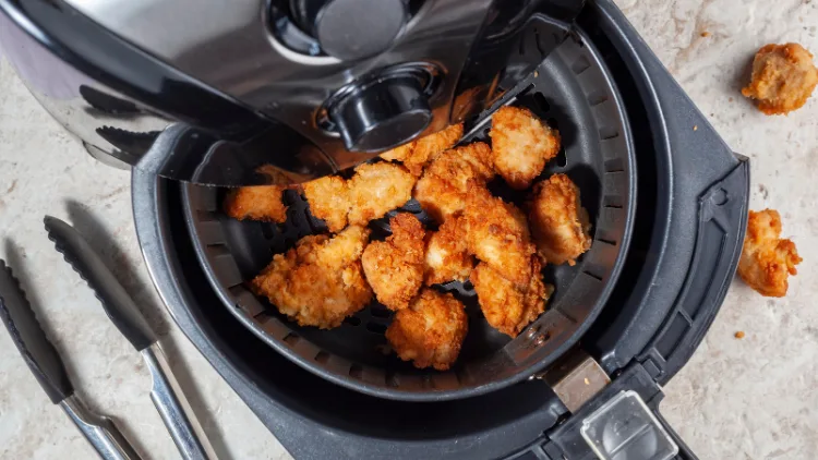 On a white concrete surface are tongs, and two pieces of fried chicken, and a black opened air fryer machine with fried chicken inside.