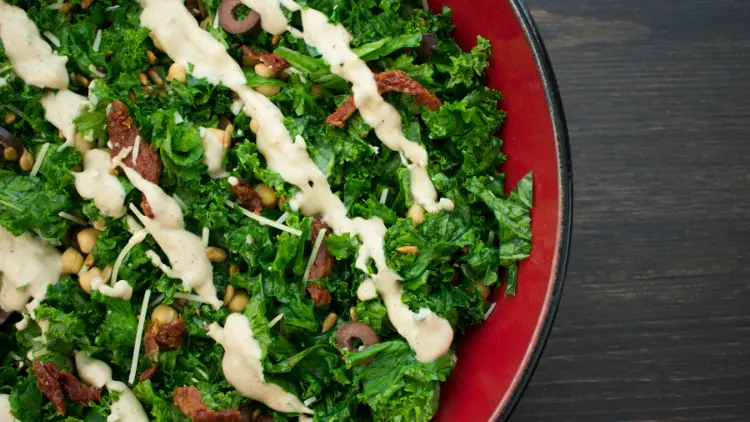 A plate of kale salad topped with a drizzle of tahini sauce, the salad features dark leafy greens, chopped vegetables, and possibly seeds or nuts, the creamy tahini sauce adds a rich, nutty flavor to the salad and complements the fresh ingredients.