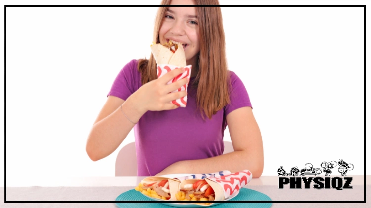 A teenage girl with red hair and a purple shirt is sitting at a table that has two paper wrappings that contain sandwiches with gyro meat inside while taking a bite from a gyro sandwich because not only is gyro keto, but it's high in protein and fat too.