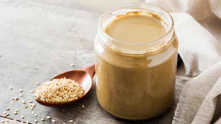 A jar of tahini sauce with a spoon resting on the side of the jar, the jar is filled with a creamy, smooth paste made from ground sesame seeds, to the side of the jar, a small pile of sesame seeds is visible, suggesting the natural ingredients used to create the tahini.