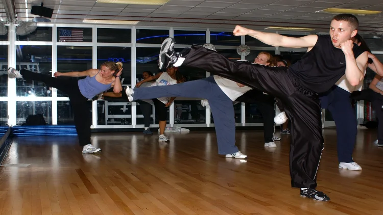 A group of students are participating in a kickboxing class, captured in mid-air as they balance on one foot with the other leg extended in front of them, they are positioned in a spacious and bright studio with large windows in the background.