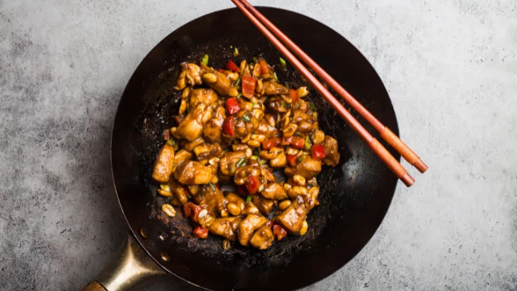 On a concrete surface os a black frying pan with Kung Pao chicken dish made with chicken meat, peppers, chili, vegetables, peanut and soy, a chopsticks can be seen placed on top of the pan.