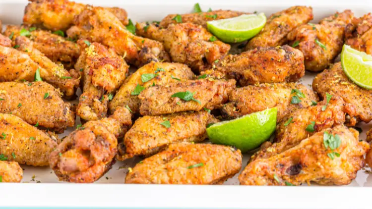 On a white rectangular platter are pieces of lemon pepper chicken wings with three lemon wedges.