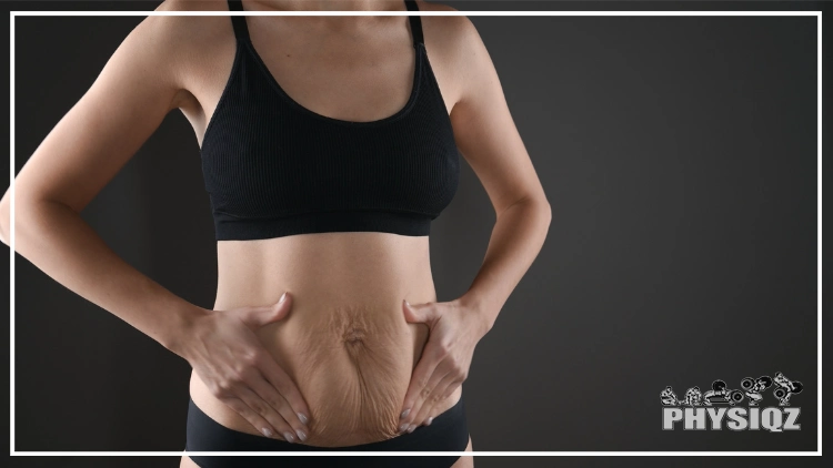 A woman wearing a black sleeveless top and black shorts is holding her stomach's loose skin after weight loss surgery in a studio against a black background.