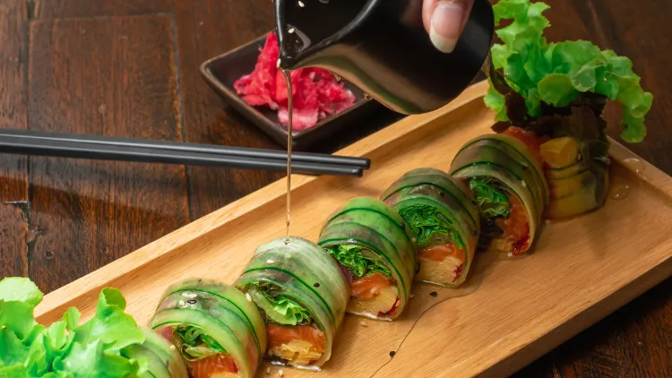 A Japanese fusion food, maki rolls without rice, made with vegetables, and seafood, served on a wooden board, with chopsticks on the side and a person is pouring a sauce on the food.