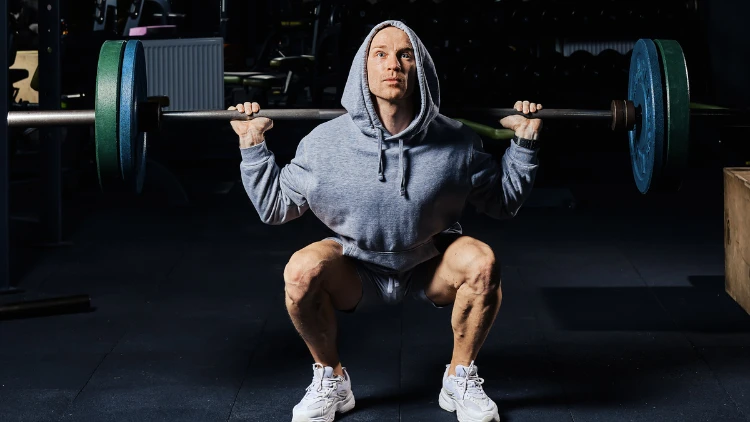 A man wearing a grey hoodie and shorts, and white shoes is performing a deadlift squat in a dimly lit gym.