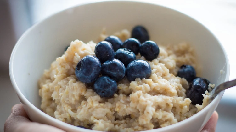 A person's hand holding a white bowl filled with oatmeal with blueberries.