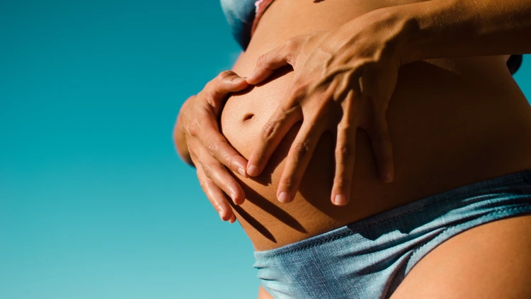 A pregnant woman wearing blue underwear in a blue background, the woman is holding her pregnant belly with both hands, forming a heart shape with her fingers around her belly button at the center.