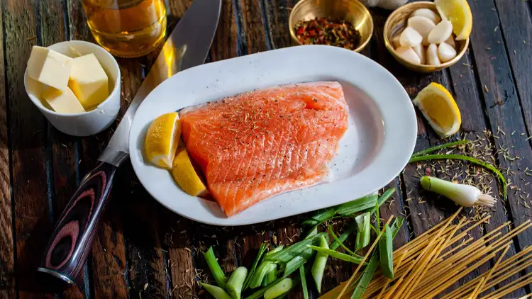 On a white oval plate is a slice of raw salmon fish seasoned with pepper and lime, and displayed on top of a wooden surface along with a cup of butter slices, raw pasta noodles, cup of cloves of garlics, and a bottle of oil.