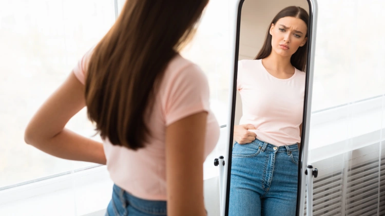 A woman wearing a light pink t-shirt, and denim pants looking at herself in the mirror while pinching her waist to measure it, and it saddens her due to weight gain.