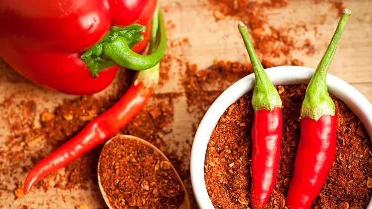 A white bowl filled with red spice powder and a pair of cayenne pepper and on the wooden surface are spills of the spice powder, a wooden spoon with spice powder, a piece of cayenne pepper, and a red bell pepper.