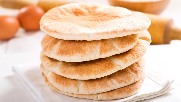 A stack of five delicious gyro pitas is on display, the pitas are soft and fluffy, with a golden brown hue from being perfectly toasted, in the background, a rolling pin can be seen.