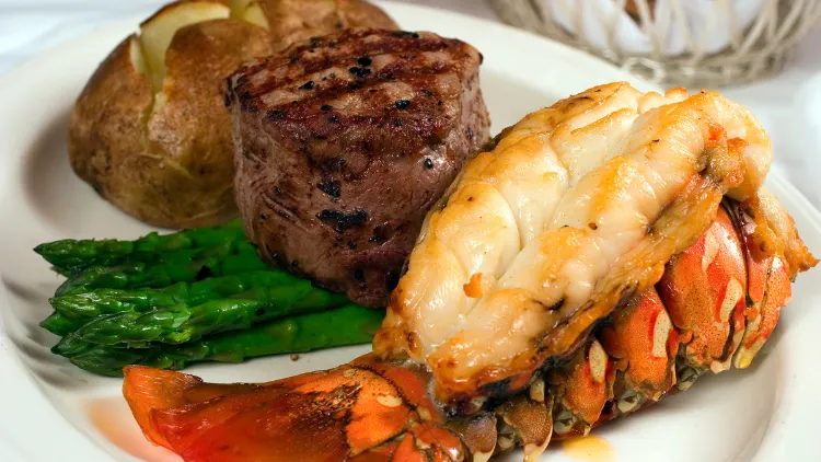 A scrumptious meal is presented on a white plate, featuring a perfectly cooked steak alongside a succulent lobster tail and a potato, below the steak, are neatly arranged asparagus, adding color and flavor to the dish.