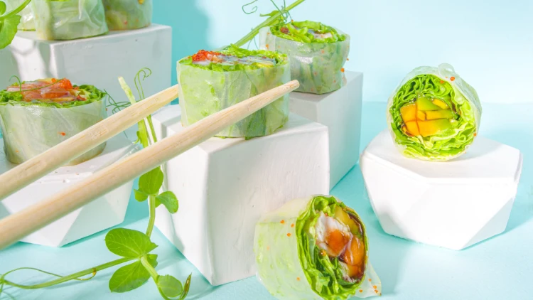 Nine sushi spring rolls without rice, but made with cucumber, kiwi, avocado, mango, and leaf vegetable wrapped with rice paper placed on white platforms with chopsticks, and everything is displayed on a light blue surface.