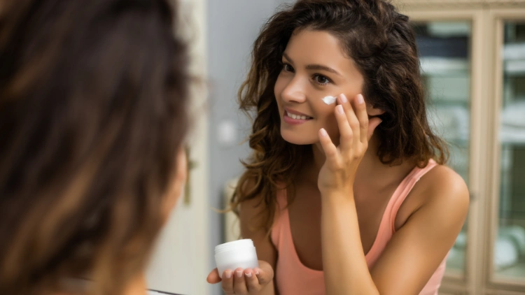A tan woman with wavy hair, wearing a pink tank top is looking at herself in the mirror while doing skin care using a face cream.