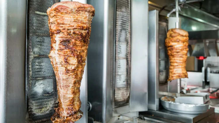 Two delicious Greek gyro meat cones rotate on a spit, cooking to perfection, with the golden-brown exterior of the meat glistening in the light, thin slices have been expertly carved off the outer layers, revealing the juicy and tender meat within.